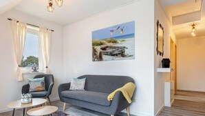 A living room with a grey sofa, a small round table, and a chair. There is a painting of birds on the wall, a window with curtains, and a wooden floor. - Vacation home Döhner Whg. 04 in Zinnowitz with balcony and 500 m to the sandy beach (Zinnowitz)