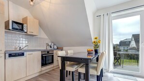 A modern kitchen with a built-in microwave, oven, and dishwasher. There is a dining table with chairs and a vase of sunflowers. The room has a large window with a view of houses outside. - Vacation home Döhner Whg. 04 in Zinnowitz with balcony and 500 m to the sandy beach (Zinnowitz)