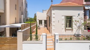 Une rue étroite, bordée de bâtiments des deux côtés, avec une entrée fermée par un portail et un petit espace extérieur aménagé avec une table et des chaises.