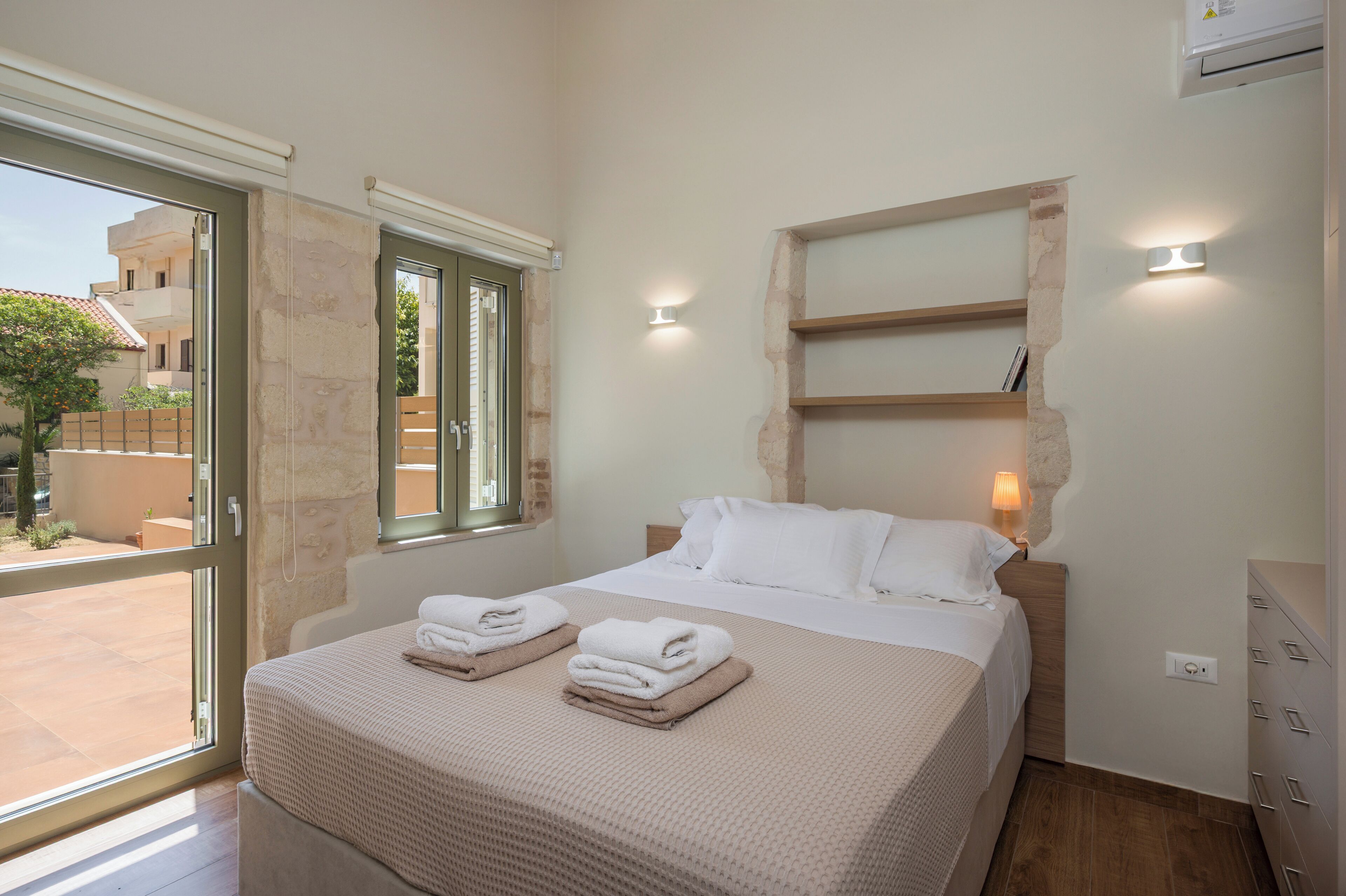 A bedroom with a bed, a window, a wall-mounted shelf, and a bedside lamp.