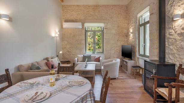 Un salon chaleureux avec un mur en pierre, un plafond en bois, une cheminée, un coin repas avec une table dressée pour deux personnes et un espace salon confortable.