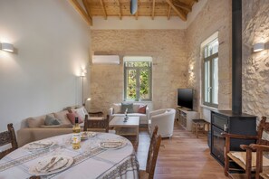 Un salon chaleureux avec un mur en pierre, un plafond en bois, une cheminée, un coin repas avec une table dressée pour deux personnes et un espace salon confortable.
