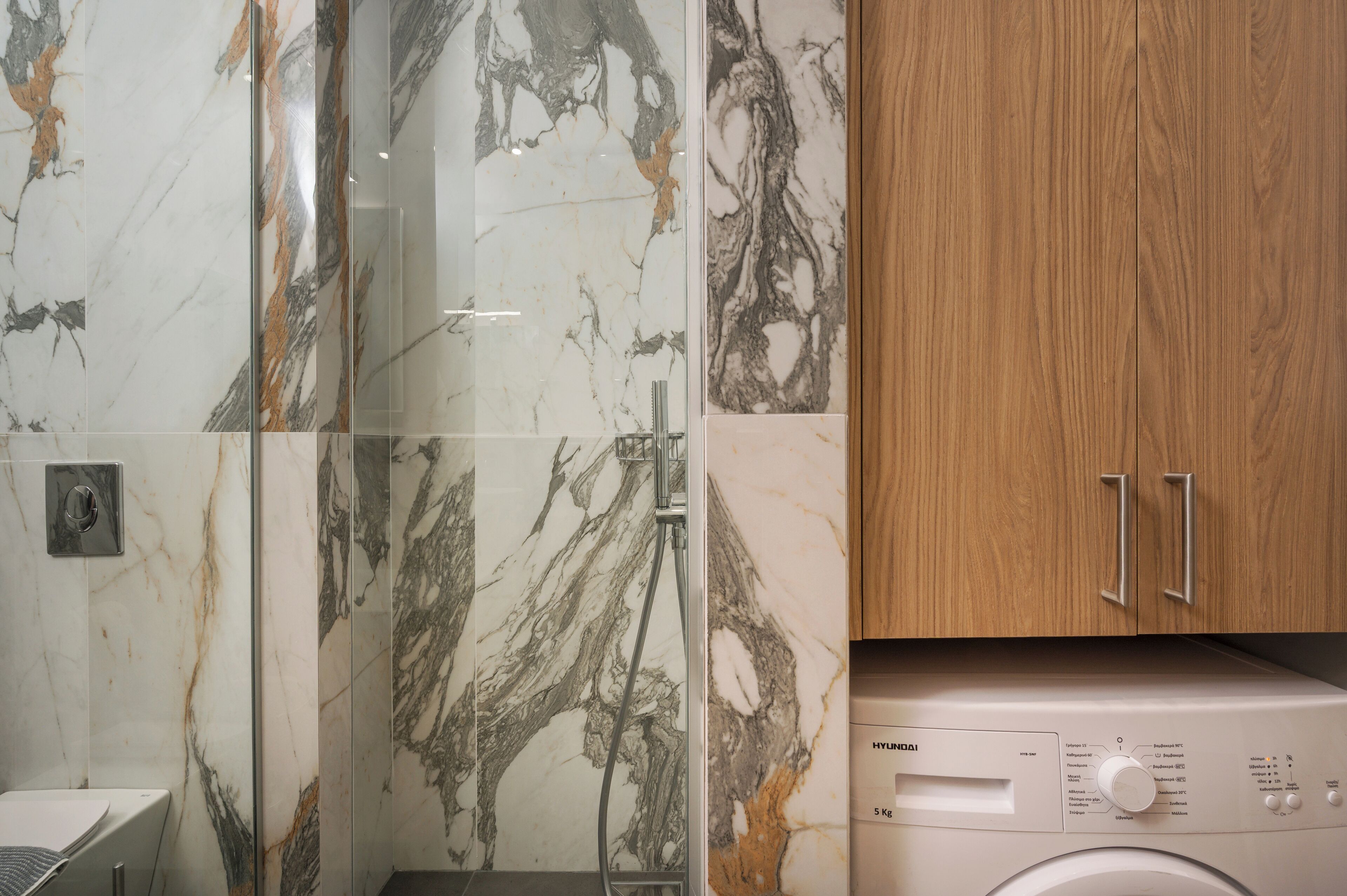 A modern bathroom with a marble shower wall, a wooden cabinet, and a front-loading washing machine.