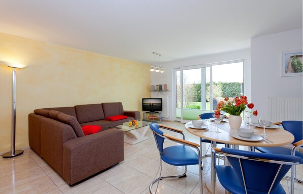 A modern living room with a brown sofa, a glass coffee table, and a dining area with blue chairs and a wooden table. - House Vincent, apartment 2 in Zinnowitz - 62 m² for up to 4 persons (Zinnowitz)