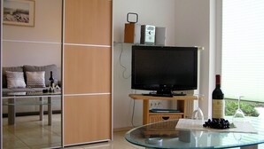 A modern living room with a glass coffee table, a flat-screen TV on a wooden stand, a sofa, and a wine bottle. - House Vincent, apartment 5 in Zinnowitz: terrace, WLAN & close to the beach (Zinnowitz)