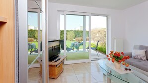 A modern living room with a glass coffee table, a sofa, a TV on a wooden stand, and a view of a garden through large sliding glass doors. - House Vincent, apartment 5 in Zinnowitz: terrace, WLAN & close to the beach (Zinnowitz)
