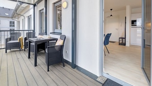 A balcony with wicker furniture and a table, a sliding glass door leading to a kitchen, and a view of neighboring buildings. - Bernsteinquartier, Whg. 02.25 in Kölpinsee - Stylish 2-room oasis near the Baltic Sea (Kölpinsee)