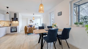 A modern kitchen with a built-in oven, a dining area with a wooden table and blue chairs, and a large window with potted plants. - Bernsteinquartier, Whg. 02.25 in Kölpinsee - Stylish 2-room oasis near the Baltic Sea (Kölpinsee)
