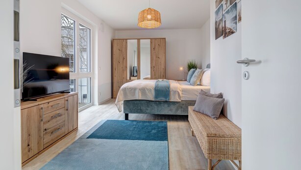 A modern bedroom with a large bed, a television, a wardrobe, and a wicker bench. - Bernsteinquartier, Whg. 02.25 in Kölpinsee - Stylish 2-room oasis near the Baltic Sea (Kölpinsee)