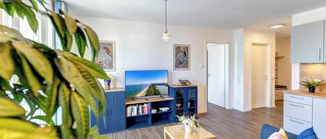 A modern living room with a sofa, coffee table, and bookshelf. There is a TV on the wall and a door leading to the kitchen.