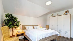 A bedroom with a wooden bed, white bedding, a potted plant, wicker chairs, a nightstand with a lamp, and a wardrobe.