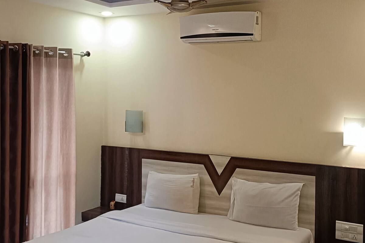 Una habitación de hotel con una cama grande, un ventilador de techo, un equipo de aire acondicionado, una mesita de noche con lámpara y una cortina.