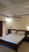 A hotel room with a large bed, a ceiling fan, an air conditioning unit, a nightstand with a lamp, and a curtain.