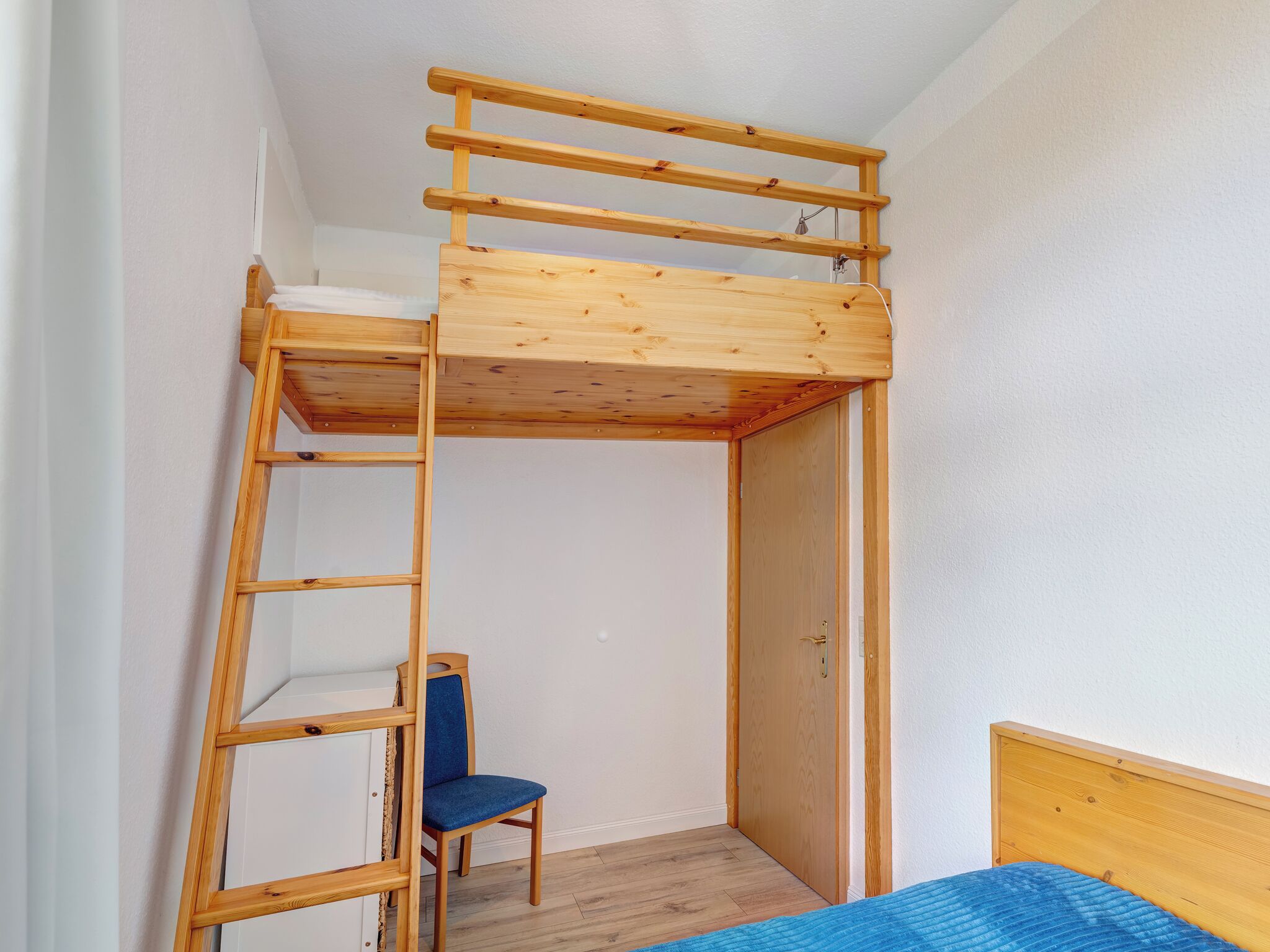 A bunk bed with a wooden ladder, a chair, and a door in a room with white walls and wooden flooring.