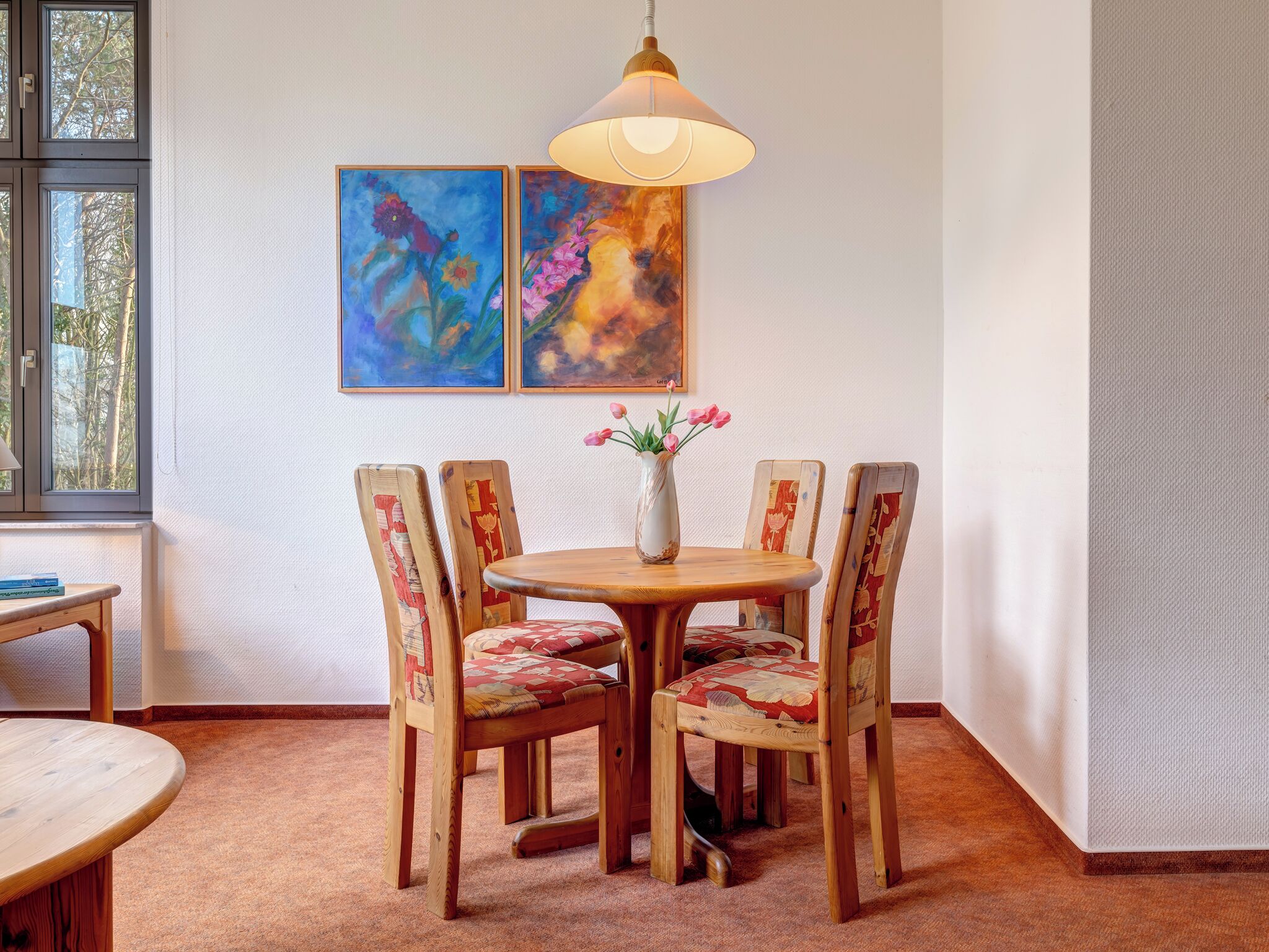 A dining area with a round wooden table, four chairs with patterned upholstery, a vase with flowers, and two abstract paintings on the wall.