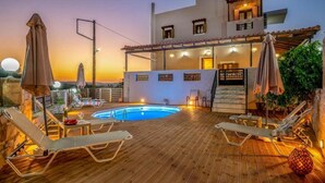 A rooftop pool area with lounge chairs and umbrellas, a pool, and a building with a terrace and lights. - Amalia Villa with Private Pool (Mylopotamos)