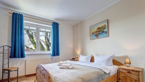 A bedroom with a wooden bed, a window with blue curtains, a radiator, a chair, a bedside table with a lamp, and a painting on the wall. - Ostseequartett, Whg. 24 Strand-im-Schuh: 55 m² for 4 persons with balcony (Zinnowitz)