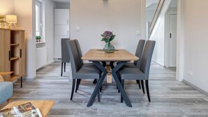 A modern dining area with a wooden table and four chairs, a vase with flowers, and a bookshelf with a lamp. - Vacation home Thorina in Bansin: 90 m² for 5, terrace & close to the beach 1 km (Bansin)