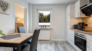 A modern kitchen with a dining area, featuring a window with a view of greenery, a built-in oven, and a coffee maker on the countertop. - Vacation home Thorina in Bansin: 90 m² for 5, terrace & close to the beach 1 km (Bansin)