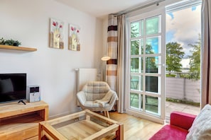 A modern living room with a flat-screen TV, a glass coffee table, a comfortable armchair, and a large sliding glass door leading to an outdoor area.