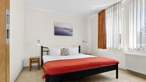 A bedroom with a bed, a nightstand, a wall-mounted lamp, a window with curtains, and a painting on the wall.