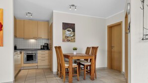 A compact kitchen with wooden cabinets, a dining table with chairs, a coffee machine, and a wall-mounted light fixture.