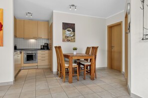 Cocina compacta con armarios de madera, una mesa de comedor con sillas, una cafetera y una lámpara fijada a la pared.