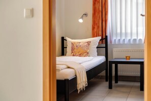 A single bed with a white mattress and a patterned pillow, a bedside table with a cup, a wall-mounted lamp, and a window with curtains.
