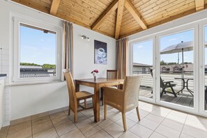 A room with a wooden ceiling, a dining table with chairs, a window with curtains, and a view of the outside.