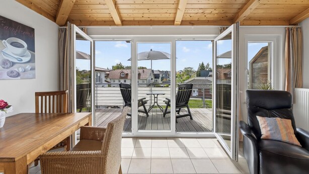 A room with a wooden ceiling, a dining table with chairs, a sofa with a cushion, and a sliding glass door leading to an outdoor patio with a table and chairs. - Am Sonnenblick Whg. 5: Apartment with balcony near the sandy beach in Heringsdorf (Heringsdorf)