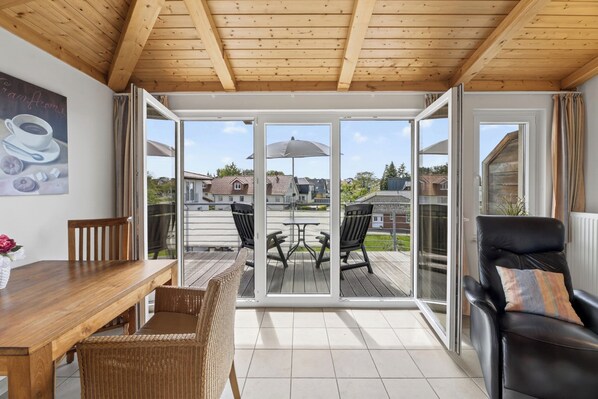A room with a wooden ceiling, a dining table with chairs, a sofa with a cushion, and a sliding glass door leading to an outdoor patio with a table and chairs.