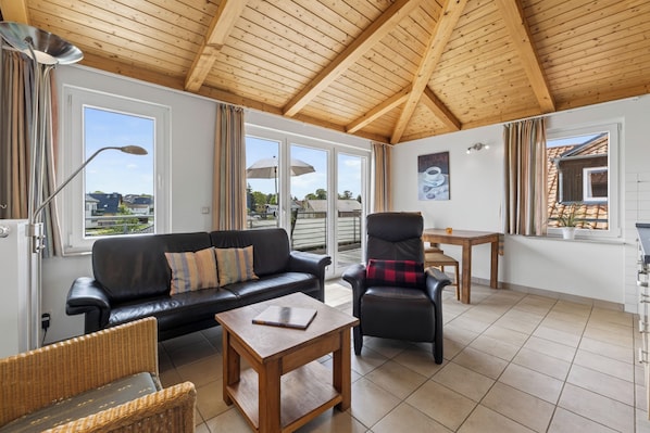 A spacious living area with a wooden ceiling, a black leather sofa, a dining table, and a chair.