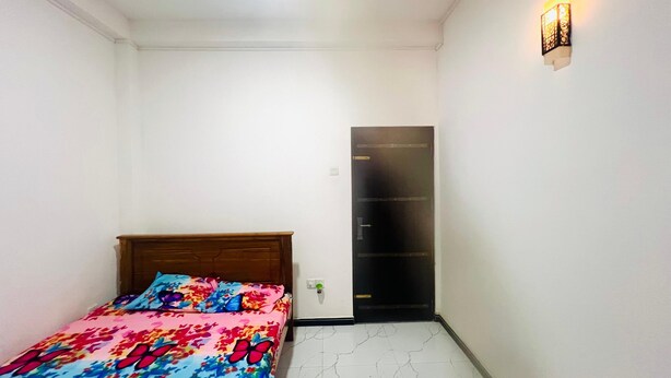 Une petite chambre bien éclairée, avec un lit simple, une armoire et un luminaire fixé au mur.
