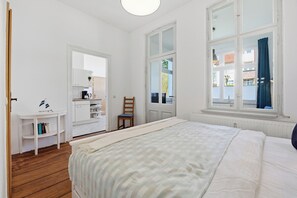 Una camera da letto con un letto, un pavimento in legno, un armadio bianco, una scrivania con libri e una finestra con tende.