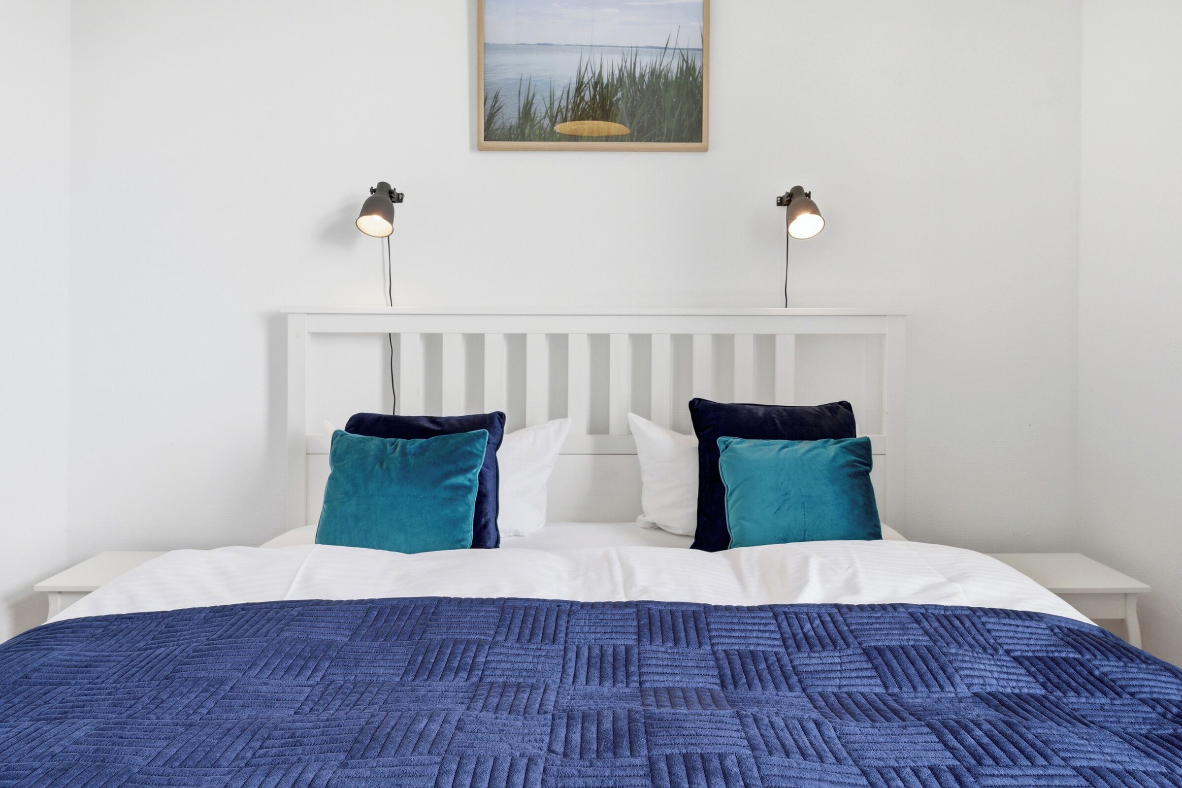 A neatly made bed with a blue textured blanket, white bedding, and teal pillows. There are two bedside lamps on either side of the bed, and a framed picture above the headboard.