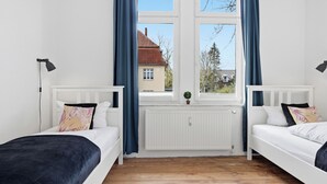 Une chambre avec deux lits simples, une fenêtre avec des rideaux, un radiateur et une petite plante.