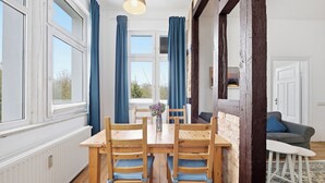 Un coin repas avec une table et des chaises en bois, un vase rempli de fleurs et une vue dégagée sur l’extérieur grâce à de grandes fenêtres ornées de rideaux bleus.