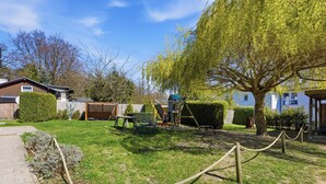 Un jardin avec une terrasse en bois, une balançoire, un espace de jeux clôturé et un grand arbre.