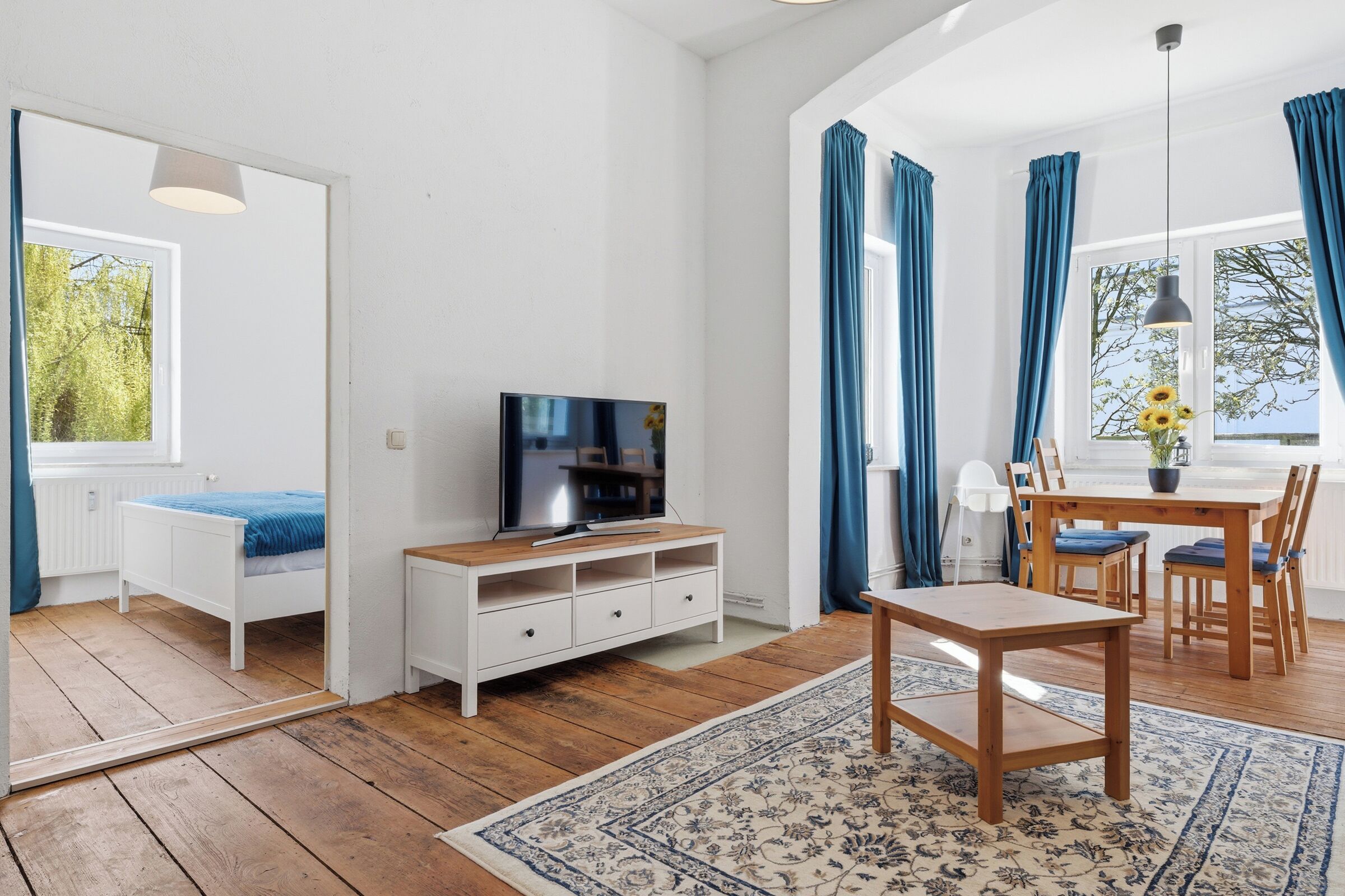 A room with a wooden floor, a TV on a white cabinet, a dining table with chairs, and a large window with curtains.