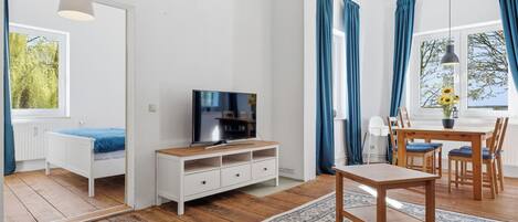 A room with a wooden floor, a TV on a white cabinet, a dining table with chairs, and a large window with curtains.
