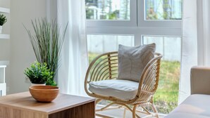 Une chaise en osier avec un coussin et un pot de plantes sur une table en bois, dans une pièce lumineuse dotée de grandes fenêtres.