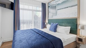 Habitación de hotel con una cama grande, un cabecero tapizado en verde, un televisor de pantalla plana montado en la pared y un cuadro de una playa.