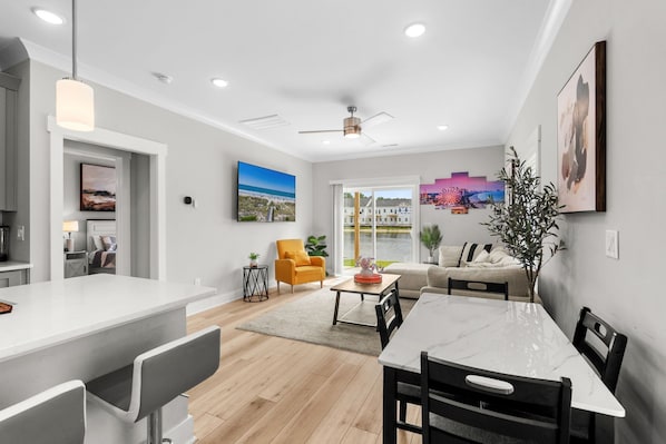A modern kitchen with a white countertop, a dining area with a marble table, and a living space with a sofa and a plant.