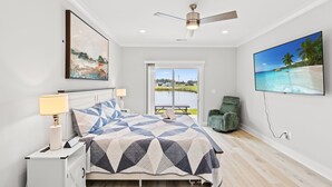 A bedroom with a bed, a nightstand, a lamp, a ceiling fan, a TV, and a window with a view of a beach. - Luxury waterfront retreat-Near golf Beach Barefoot (Longs)