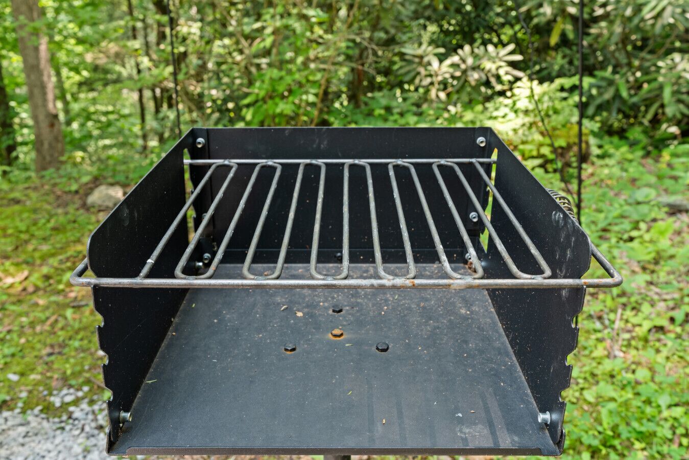 Una barbacoa negra amb una reixa metàl·lica, situada davant d'un paisatge natural de boscos i vegetació.