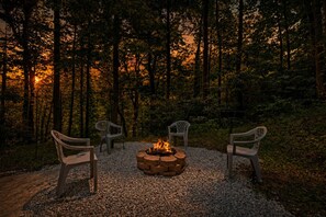 A fire pit with three chairs arranged around it in a forest setting.