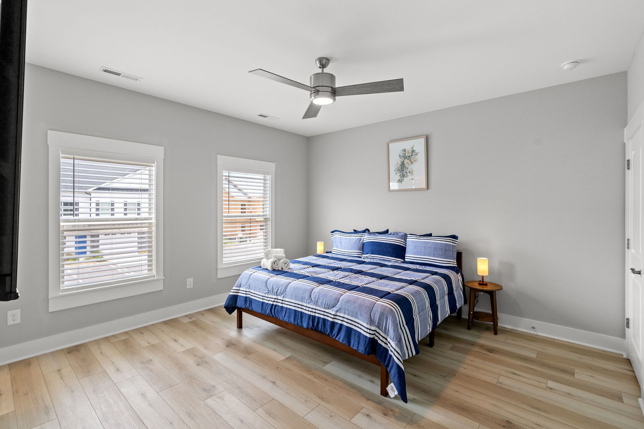 Une chambre à coucher comprenant un lit, deux tables de chevet, un ventilateur de plafond et de grandes fenêtres équipées de stores.