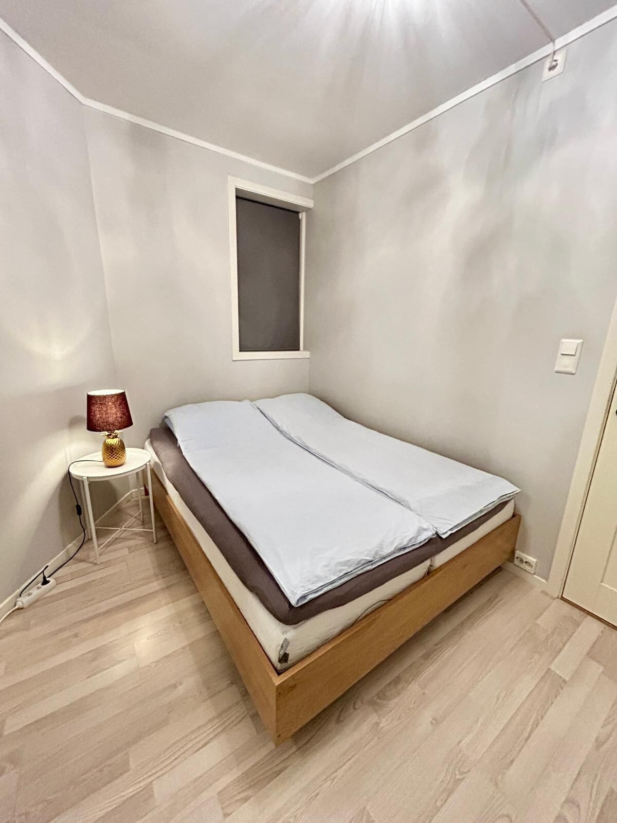 Un dormitorio pequeño y ordenado con una cama individual, una mesita con una lámpara y una ventana.