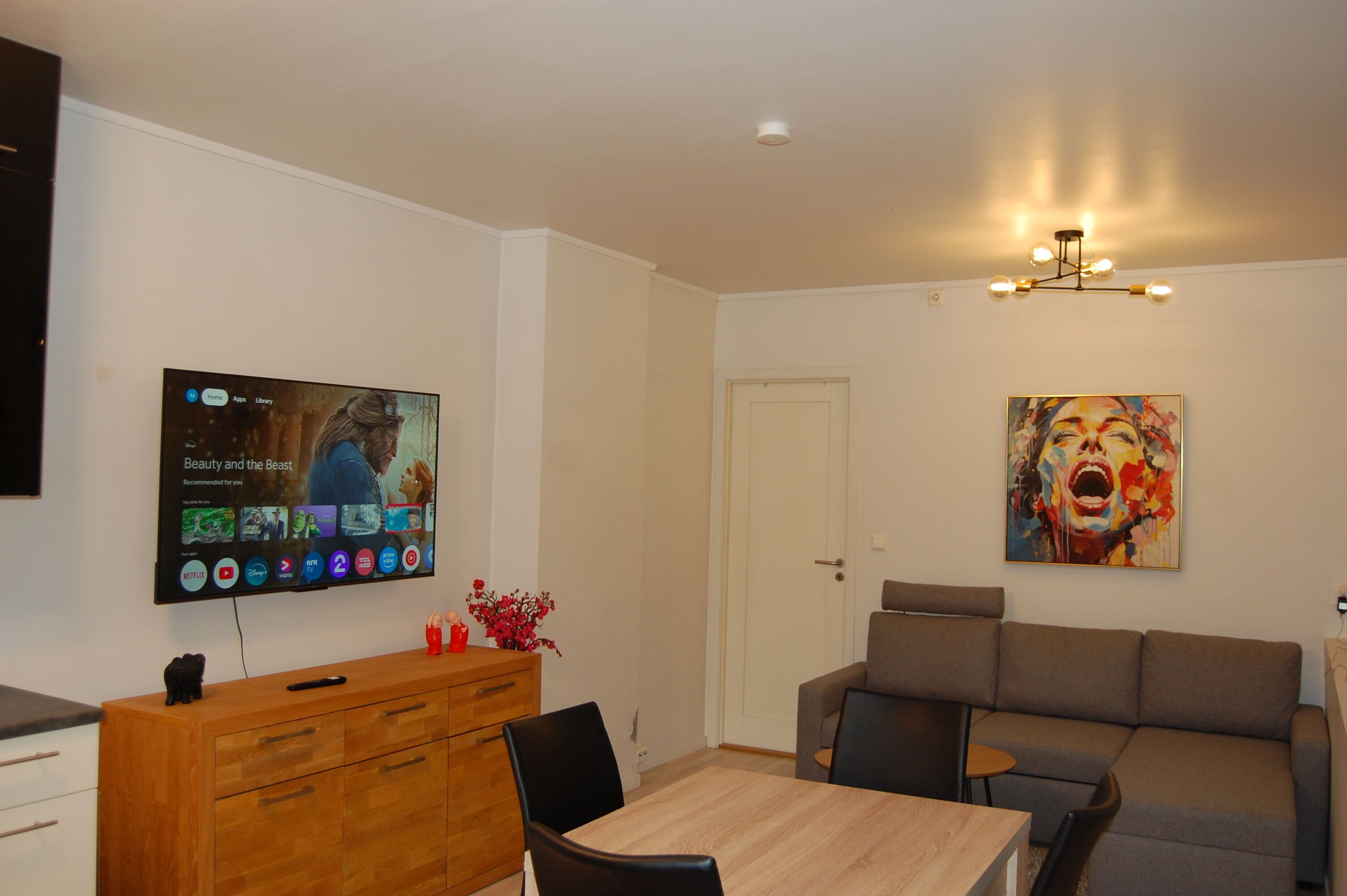 A modern living room with a flat-screen TV mounted on the wall, a wooden cabinet, a grey sofa, and a painting.