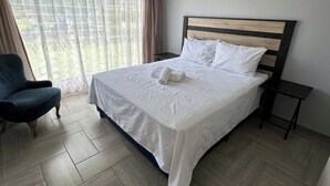 Un lit bien fait, avec du linge de lit blanc et des oreillers, un fauteuil bleu foncé et une petite table de chevet.